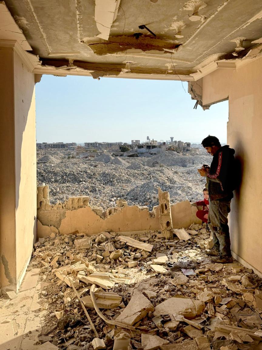 HLP GIZ Blick aus einem zerstörten Gebäude mit herabhängender Decke auf ein weitläufiges Trümmerfeld, im Vordergrund ein stehender Mann und ein Kind in der Ecke.