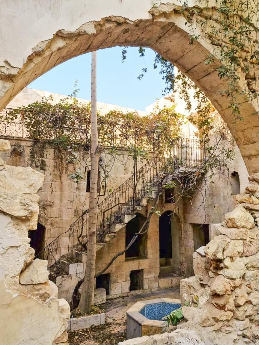 HLP GIZ Zerfallener Innenhof mit Steinbogen, verwitterter Steintreppe und einem Brunnen, teilweise überwachsen von Pflanzen.