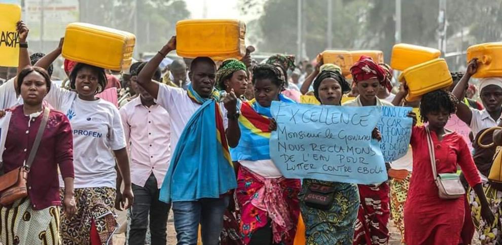 Eine Gruppe von Menschen marschiert auf einer staubigen Straße im Kongo, viele tragen gelbe Wasserkanister auf dem Kopf. Eine Person hält ein Plakat hoch, auf dem auf Französisch steht:  Exzellenz, Herr Gouverneur, wir fordern sauberes Wasser, um gegen Ebola zu kämpfen. Einige tragen traditionelle, bunte Kleidung, andere einfache Alltagskleidung.